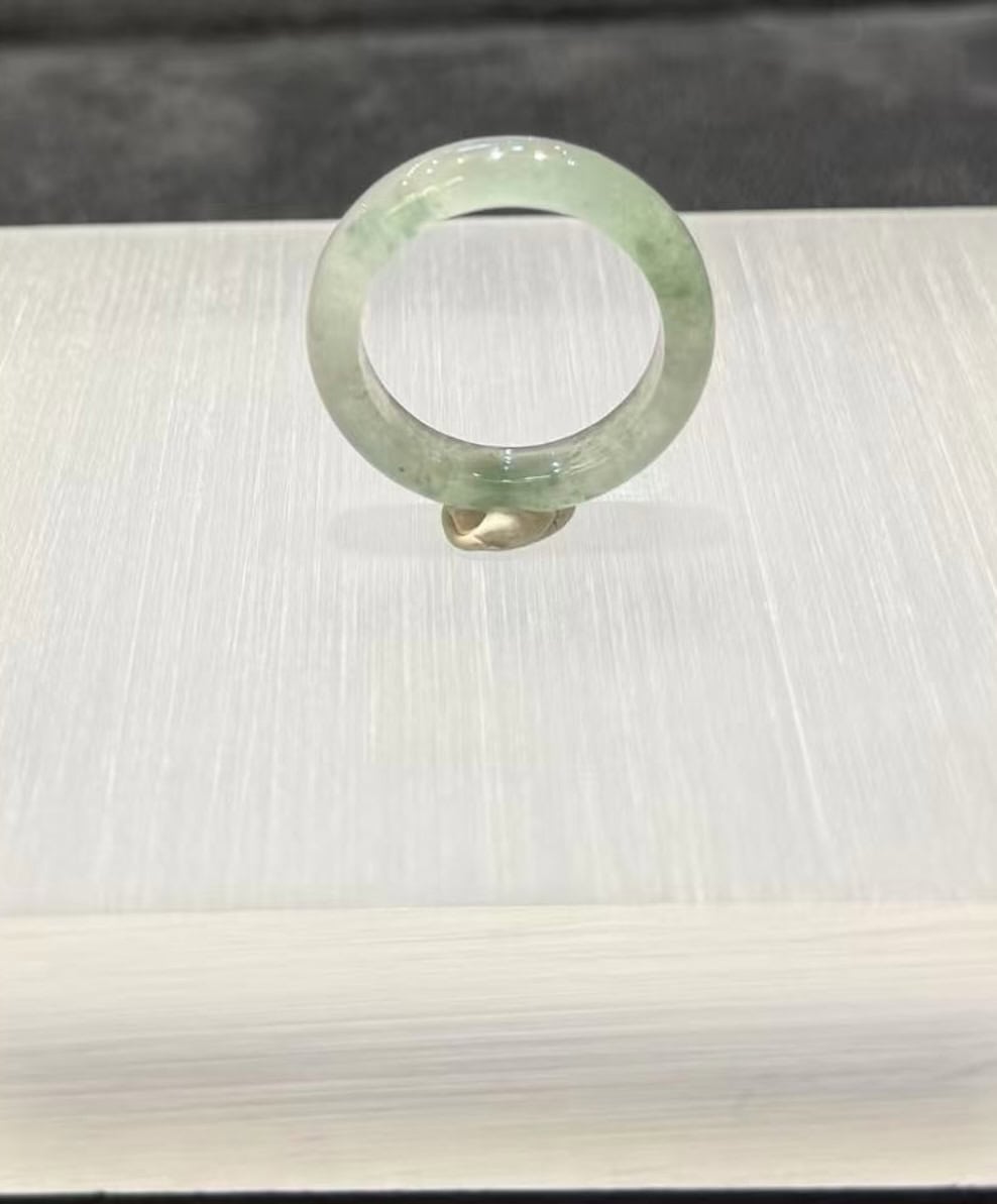 Icy jade ring carved from a single piece of translucent jadeite, with soft green floating-flower patches drifting through a clear, glassy band, shown on a white display stand