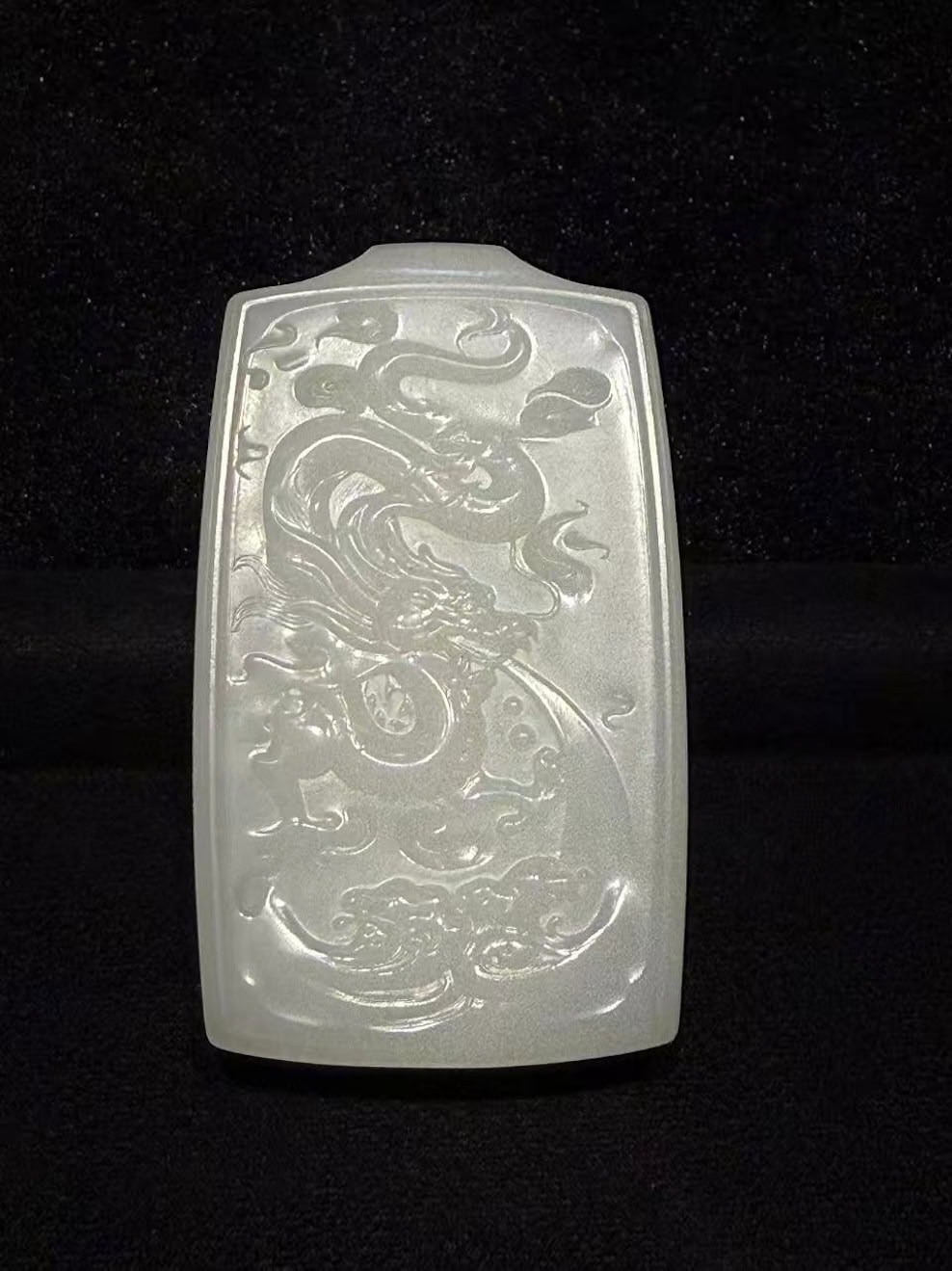Creamy white nephrite jade pendant hand-carved with a dragon splashing through rolling waves in low relief on a traditional rectangular pai-zi plaque, displayed on black velvet