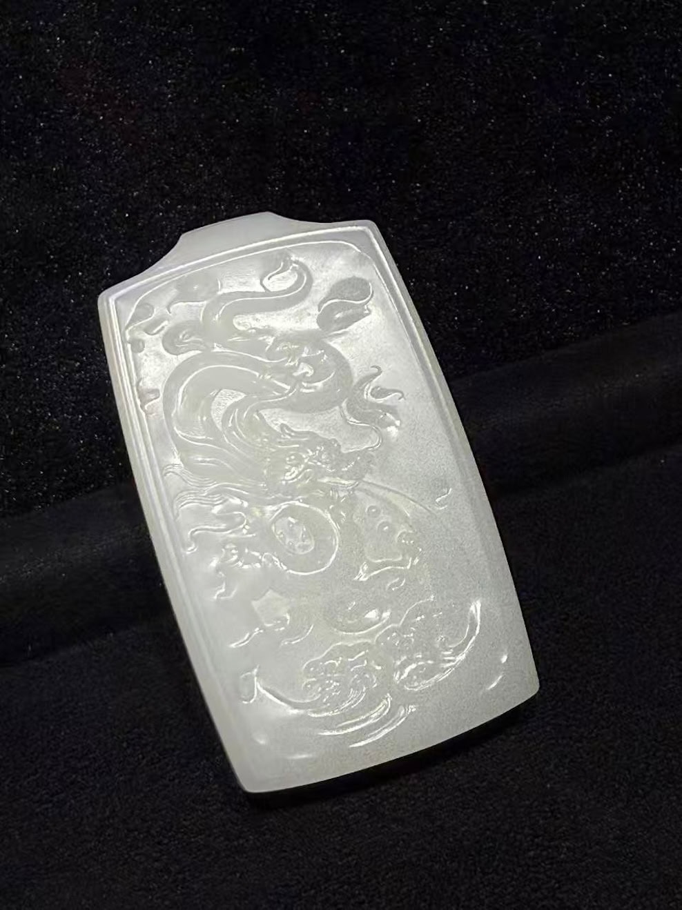 Creamy white nephrite jade pendant hand-carved with a dragon splashing through rolling waves in low relief on a traditional rectangular pai-zi plaque, displayed on black velvet