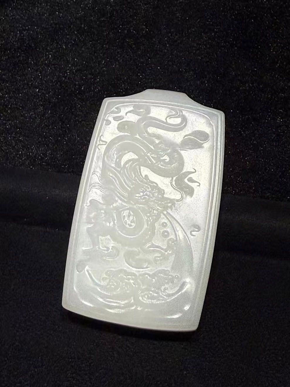 Creamy white nephrite jade pendant hand-carved with a dragon splashing through rolling waves in low relief on a traditional rectangular pai-zi plaque, displayed on black velvet