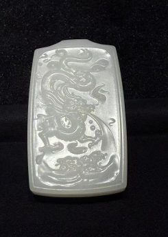 Creamy white nephrite jade pendant hand-carved with a dragon splashing through rolling waves in low relief on a traditional rectangular pai-zi plaque, displayed on black velvet