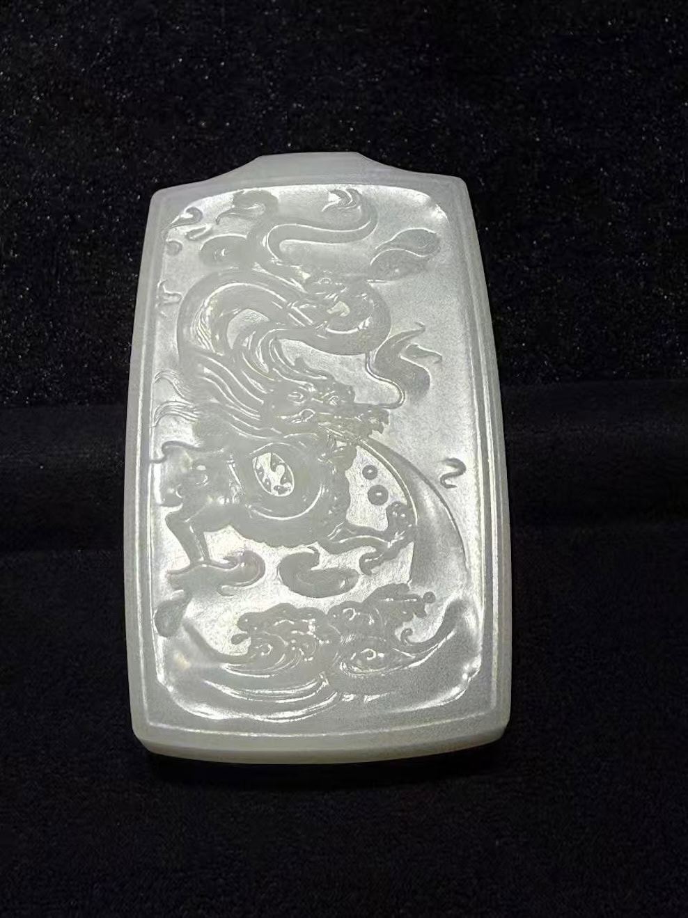 Creamy white nephrite jade pendant hand-carved with a dragon splashing through rolling waves in low relief on a traditional rectangular pai-zi plaque, displayed on black velvet