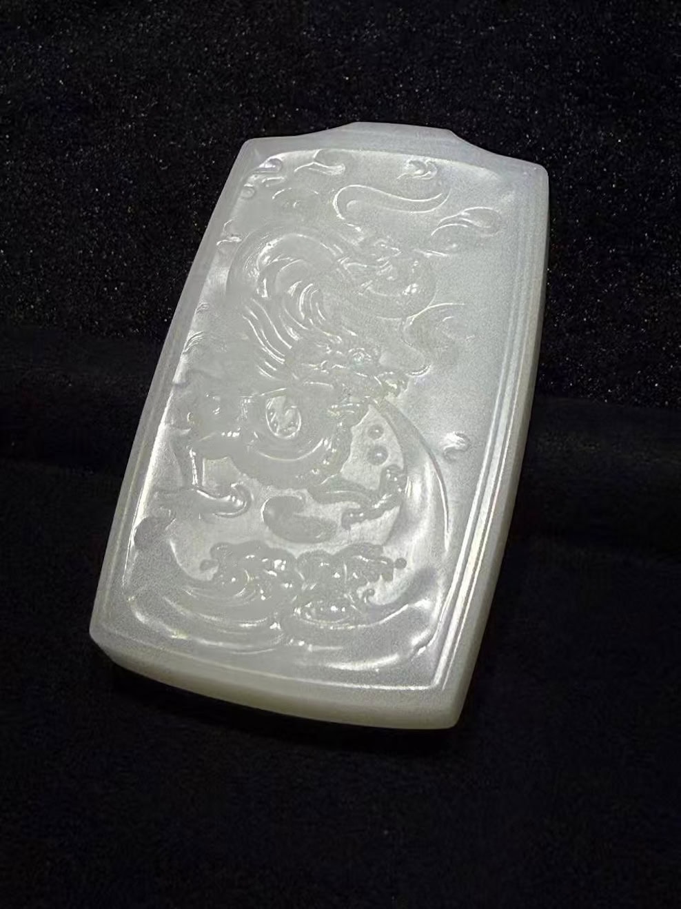 Creamy white nephrite jade pendant hand-carved with a dragon splashing through rolling waves in low relief on a traditional rectangular pai-zi plaque, displayed on black velvet
