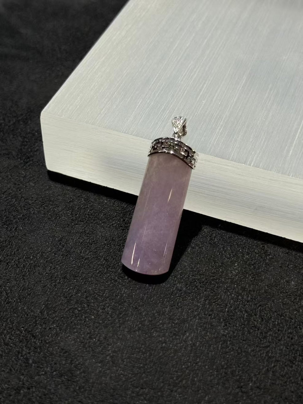 Lavender jade pendant shaped as a smooth uncarved jadeite cylinder, set in 18k white gold with a diamond bail and fretwork cap, displayed on a white stand