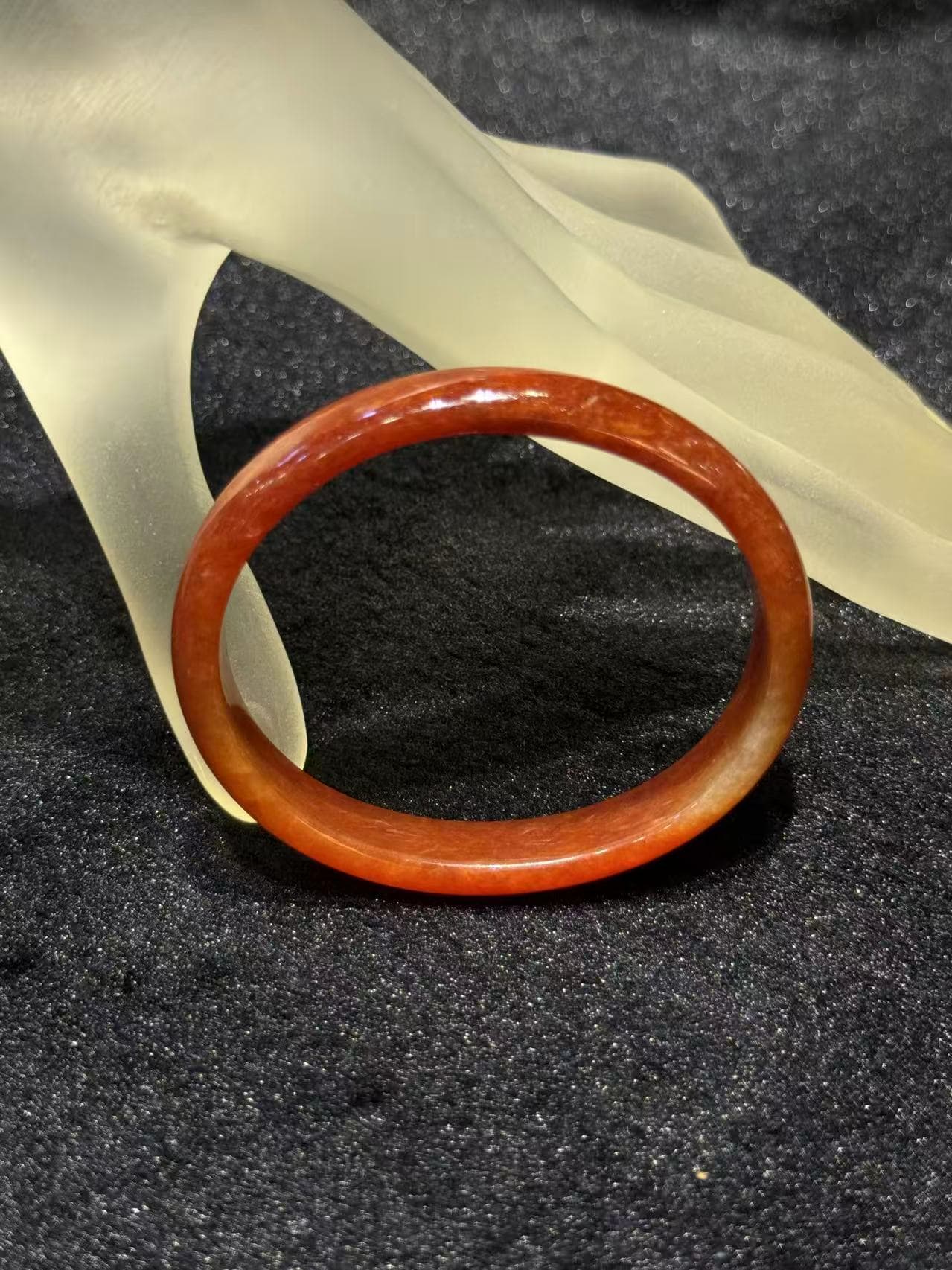 Natural red jade bangle in a warm persimmon tone with subtle internal texture, resting on a frosted hand stand against black velvet