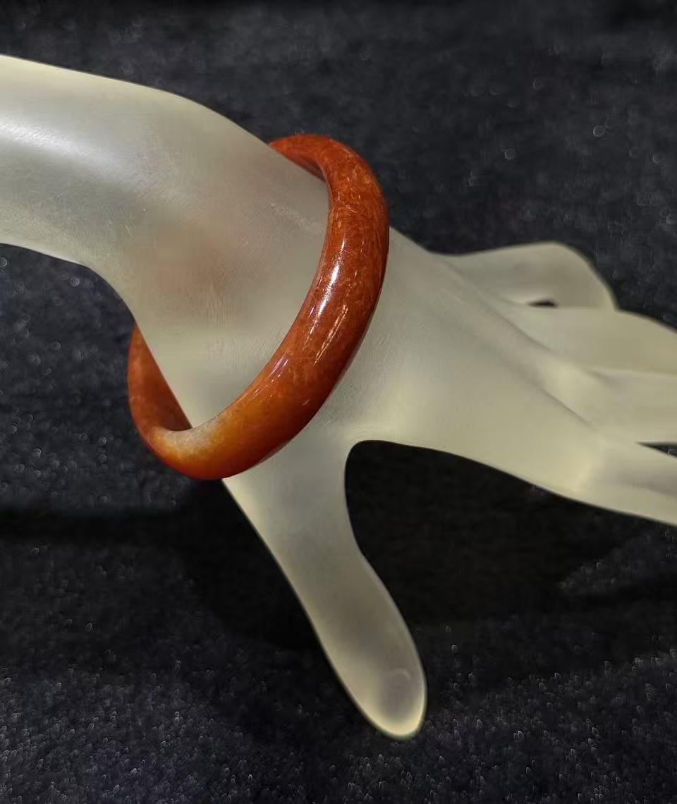 Natural red jade bangle in a warm persimmon tone with subtle internal texture, resting on a frosted hand stand against black velvet