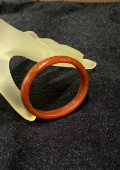 Natural red jade bangle in a warm persimmon tone with subtle internal texture, resting on a frosted hand stand against black velvet