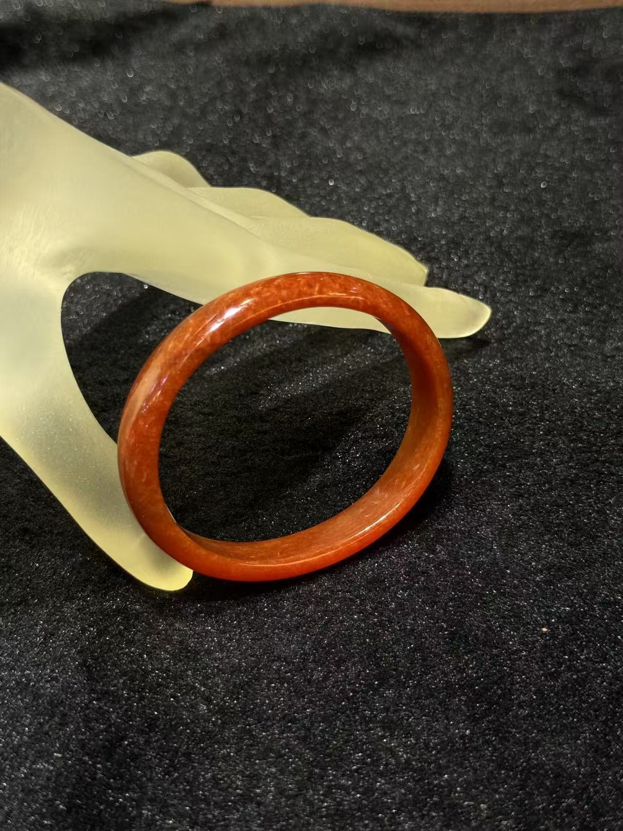 Natural red jade bangle in a warm persimmon tone with subtle internal texture, resting on a frosted hand stand against black velvet