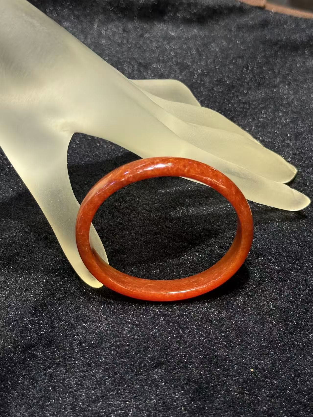 Natural red jade bangle in a warm persimmon tone with subtle internal texture, resting on a frosted hand stand against black velvet