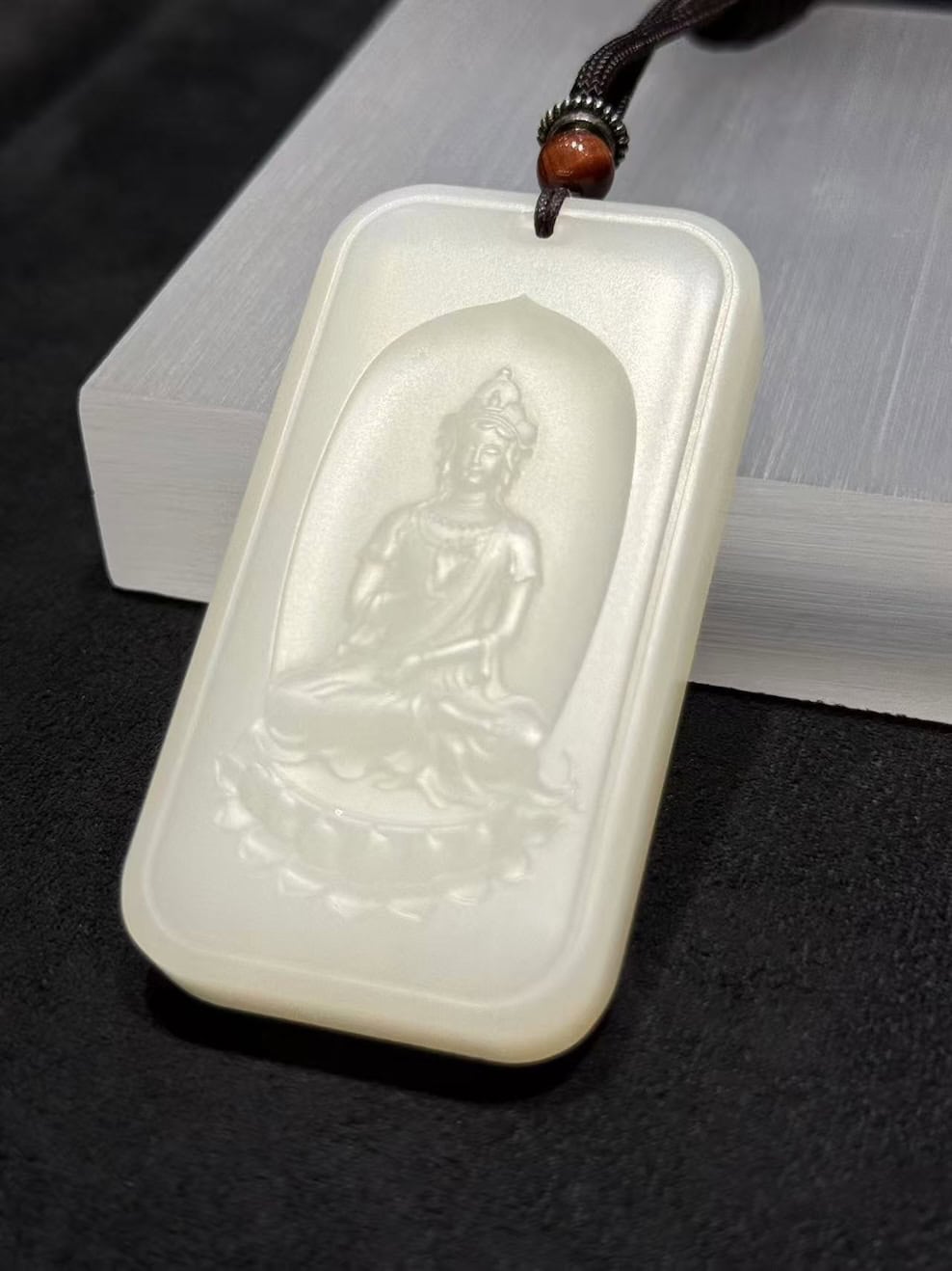 Guanyin nephrite pendant carved from soft white colour, showing the seated Bodhisattva on a lotus within a rectangular pai-zi plaque with a cloud-pattern border, on a black cord