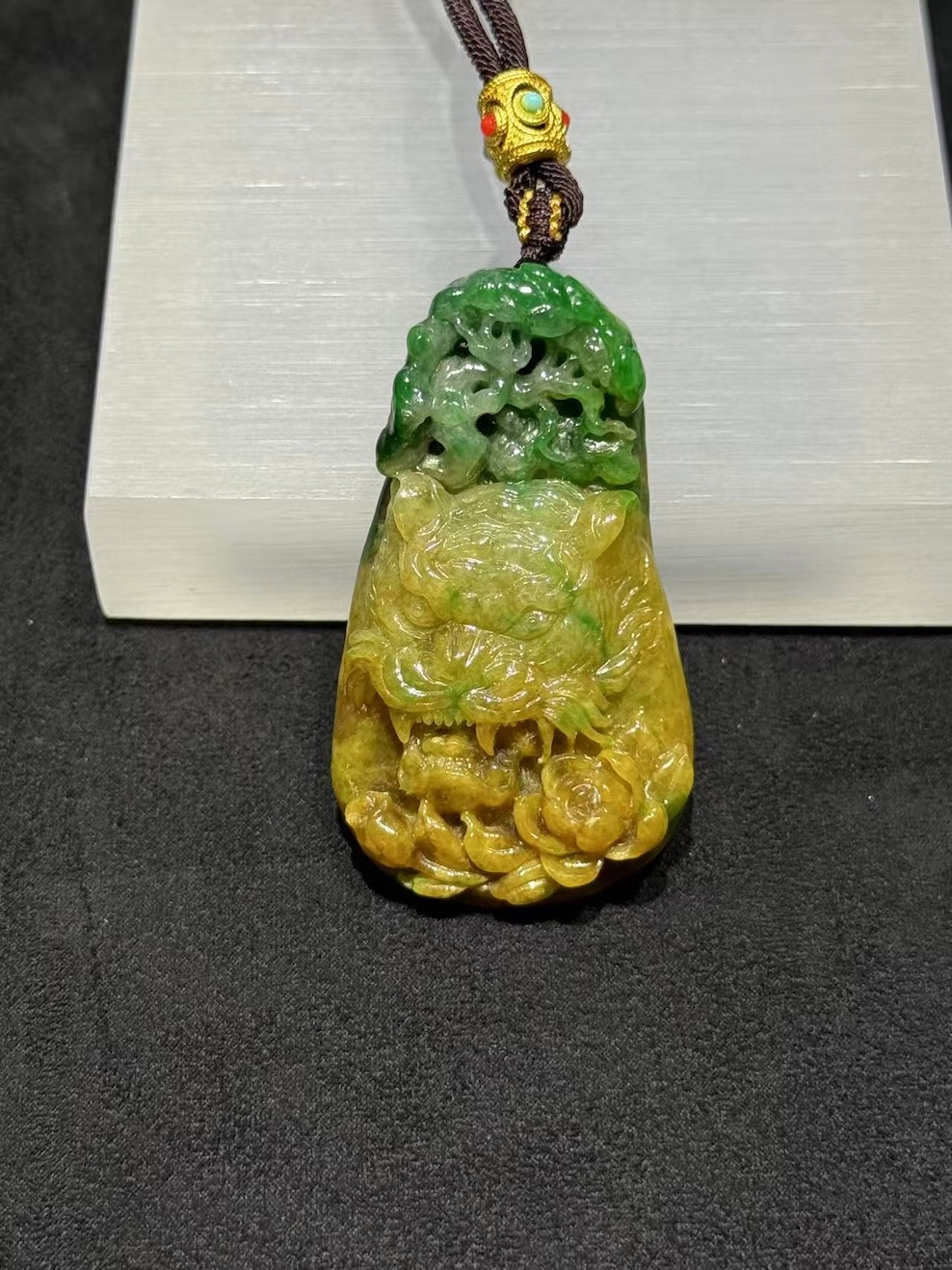 Tiger jade pendant carved from translucent yellow and green jadeite, with a detailed tiger head at the centre, a lotus blossom at the base, and openwork scrollwork above, hung on a brown rope with a small brass bead