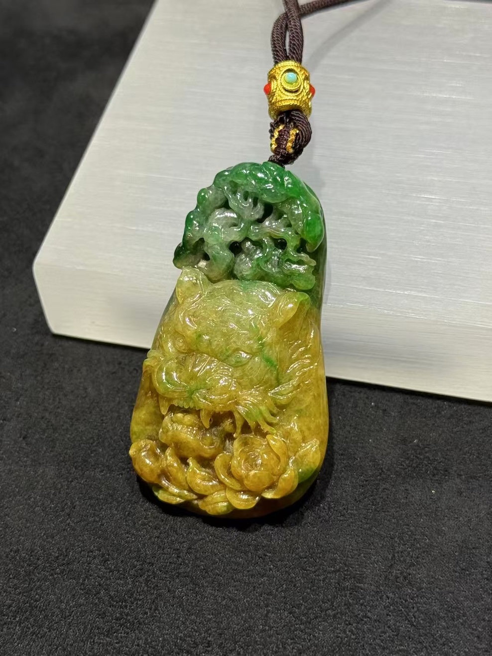 Tiger jade pendant carved from translucent yellow and green jadeite, with a detailed tiger head at the centre, a lotus blossom at the base, and openwork scrollwork above, hung on a brown rope with a small brass bead