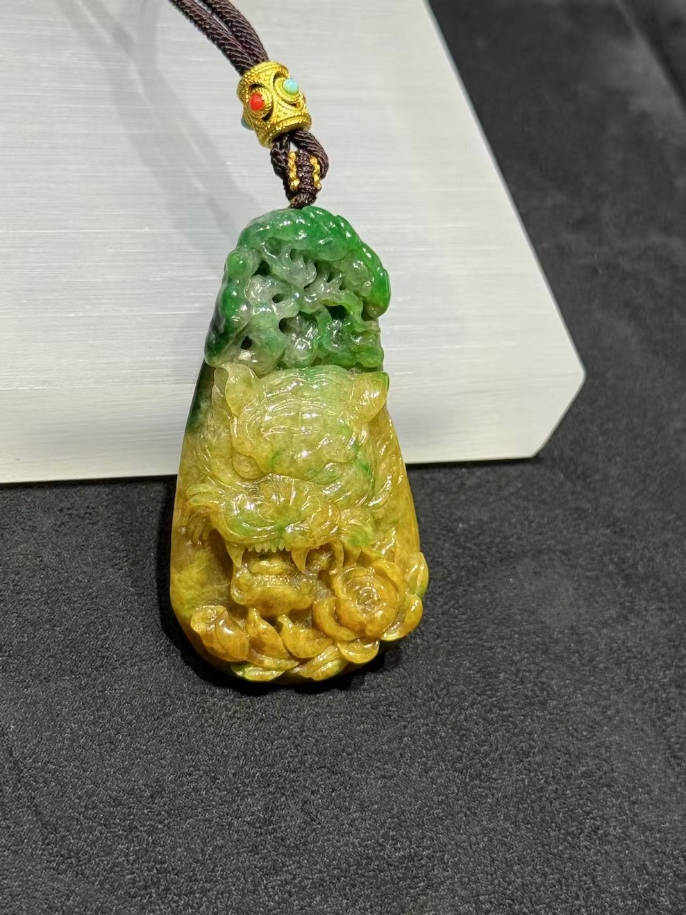 Tiger jade pendant carved from translucent yellow and green jadeite, with a detailed tiger head at the centre, a lotus blossom at the base, and openwork scrollwork above, hung on a brown rope with a small brass bead