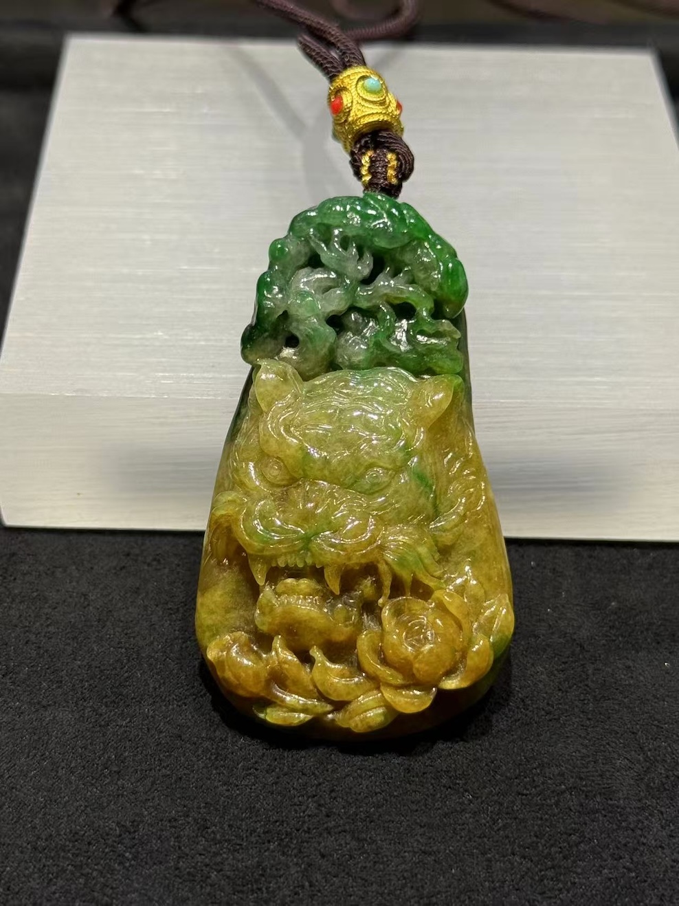 Tiger jade pendant carved from translucent yellow and green jadeite, with a detailed tiger head at the centre, a lotus blossom at the base, and openwork scrollwork above, hung on a brown rope with a small brass bead