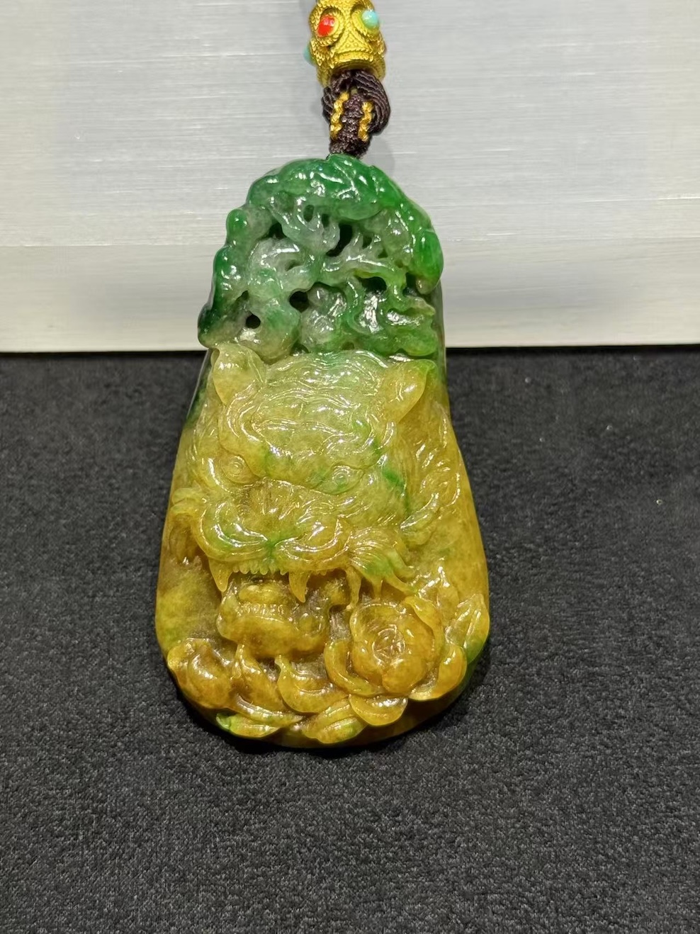 Tiger jade pendant carved from translucent yellow and green jadeite, with a detailed tiger head at the centre, a lotus blossom at the base, and openwork scrollwork above, hung on a brown rope with a small brass bead