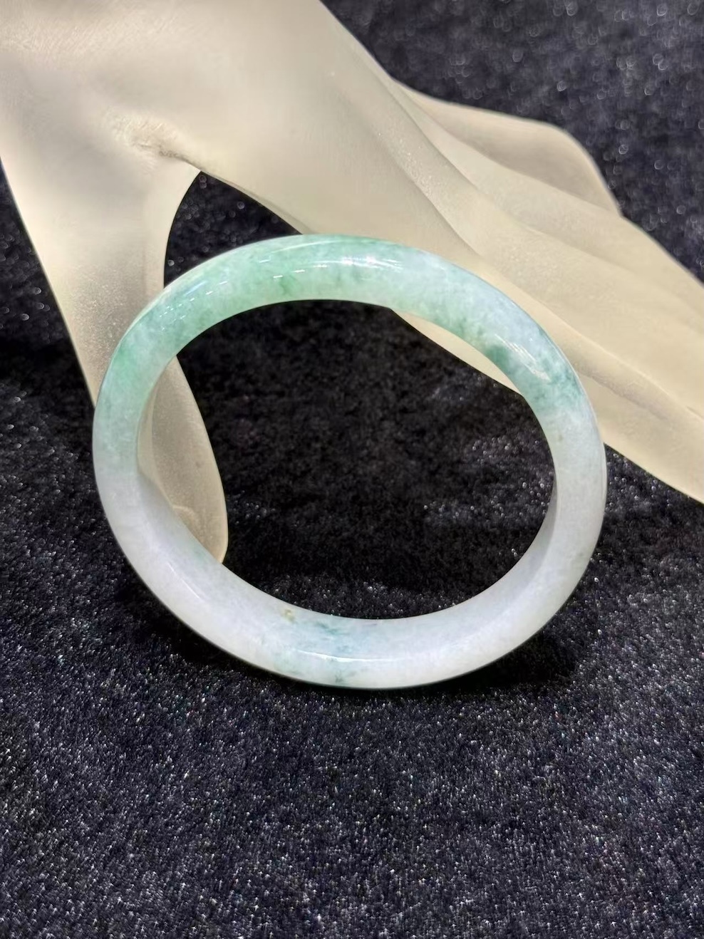 Light apple green jadeite bangle in an oval shape, with a translucent white base and soft apple green and faint bluish-green tones drifting across the ring