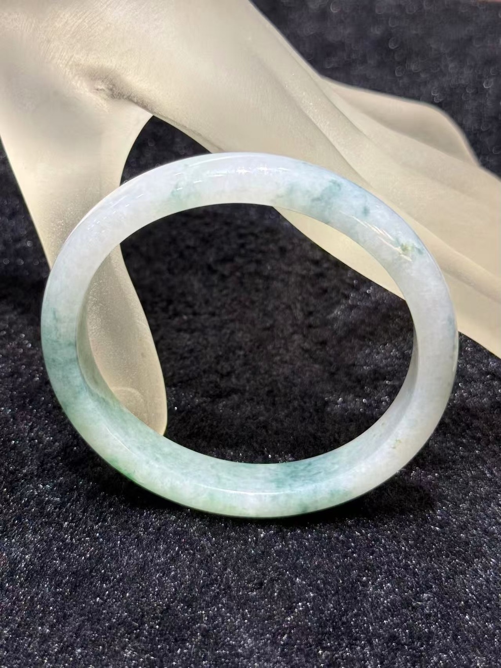 Light apple green jadeite bangle in an oval shape, with a translucent white base and soft apple green and faint bluish-green tones drifting across the ring
