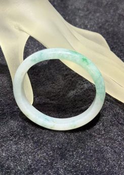 Light apple green jadeite bangle in an oval shape, with a translucent white base and soft apple green and faint bluish-green tones drifting across the ring