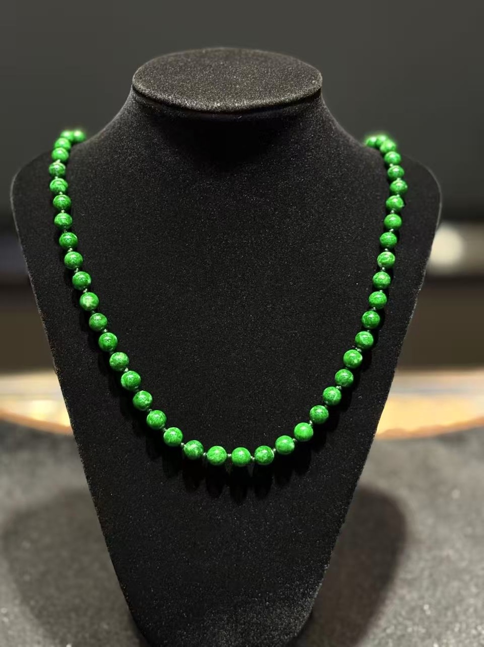 Green jade necklace strung with round vibrant green jadeite beads in an even, saturated tone, shown on a black necklace display bust
