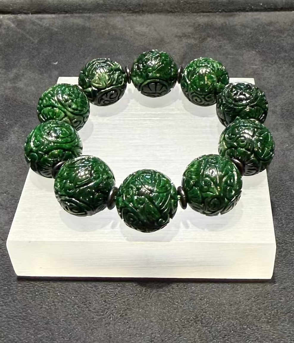 Carved jadeite bracelet strung with large dark green round beads, each cut with deep scrollwork and floral patterns, separated by small dark spacer beads