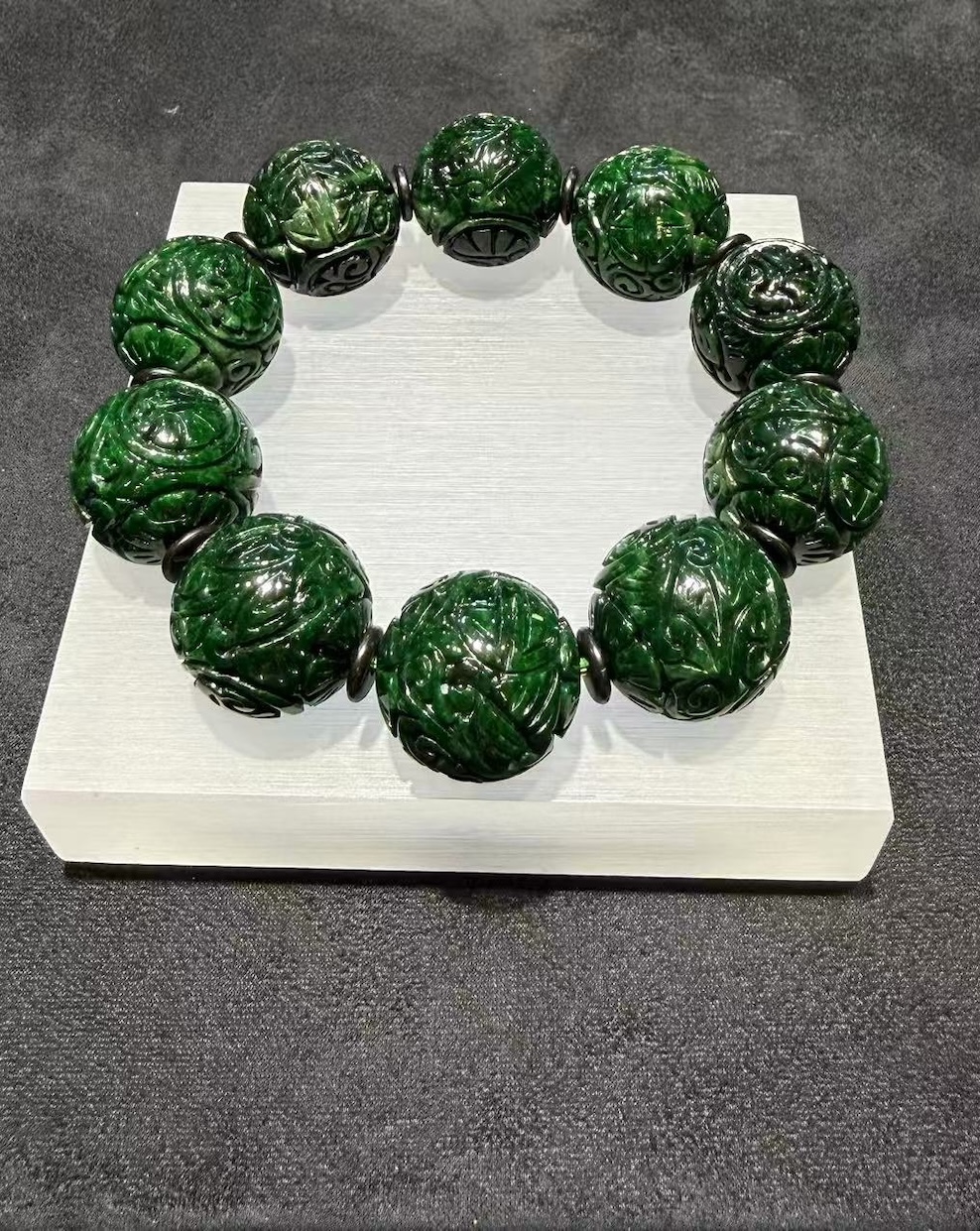 Carved jadeite bracelet strung with large dark green round beads, each cut with deep scrollwork and floral patterns, separated by small dark spacer beads