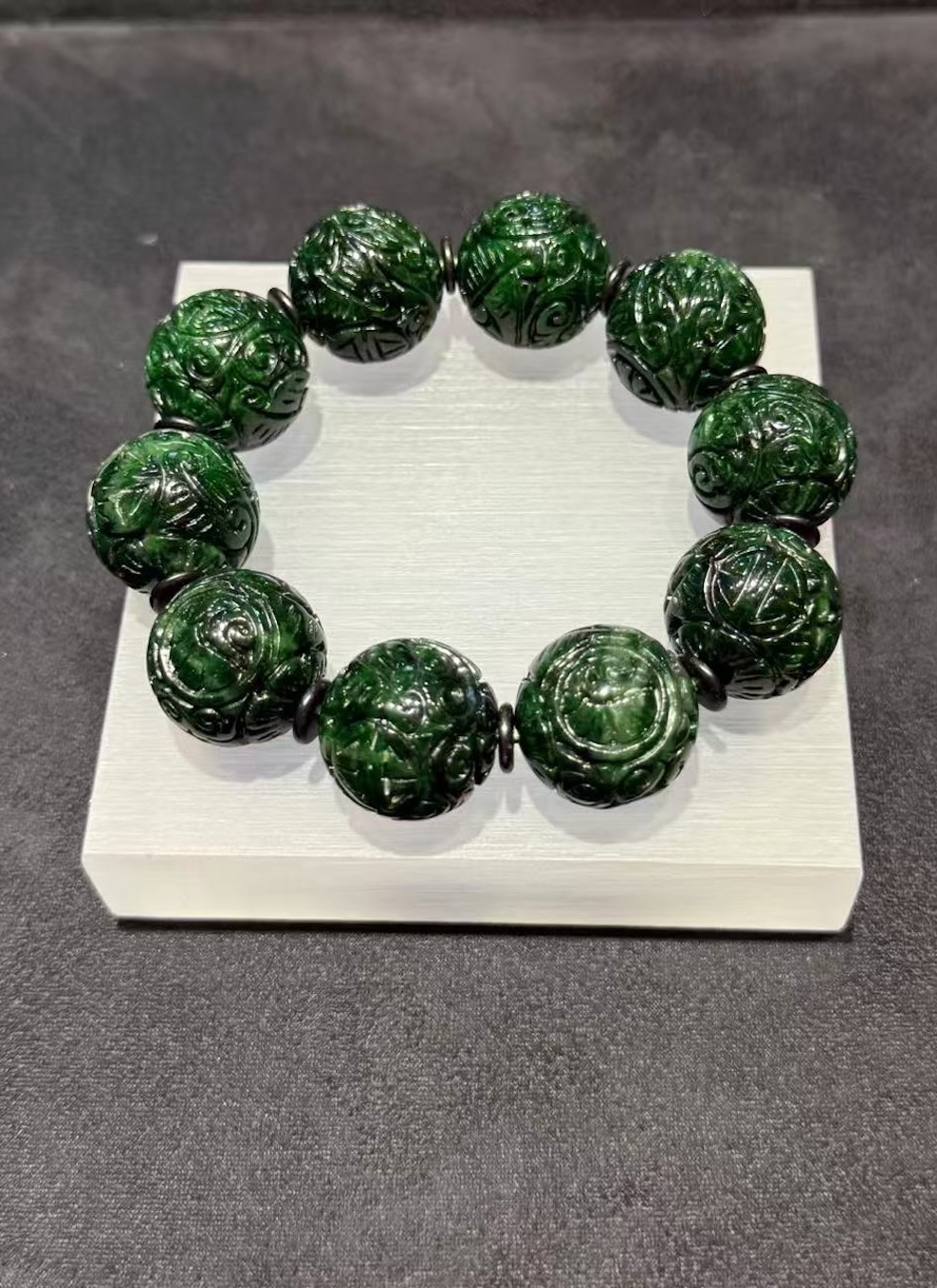 Carved jadeite bracelet strung with large dark green round beads, each cut with deep scrollwork and floral patterns, separated by small dark spacer beads