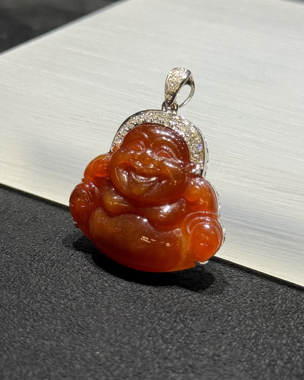 Laughing buddha jade pendant carved from translucent brownish red jadeite showing a smiling Buddha with a round belly, set in an 18k white gold bail with small diamonds