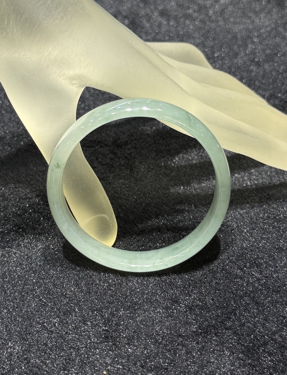 Translucent jadeite bangle in an icy, near-colourless body with a very faint green tint and small darker green flecks drifting through the ring