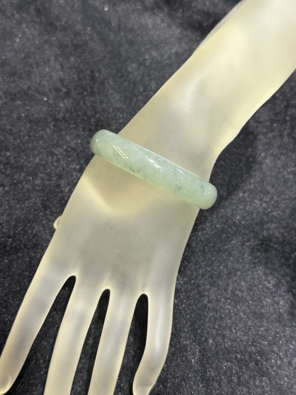 Translucent jadeite bangle in an icy, near-colourless body with a very faint green tint and small darker green flecks drifting through the ring