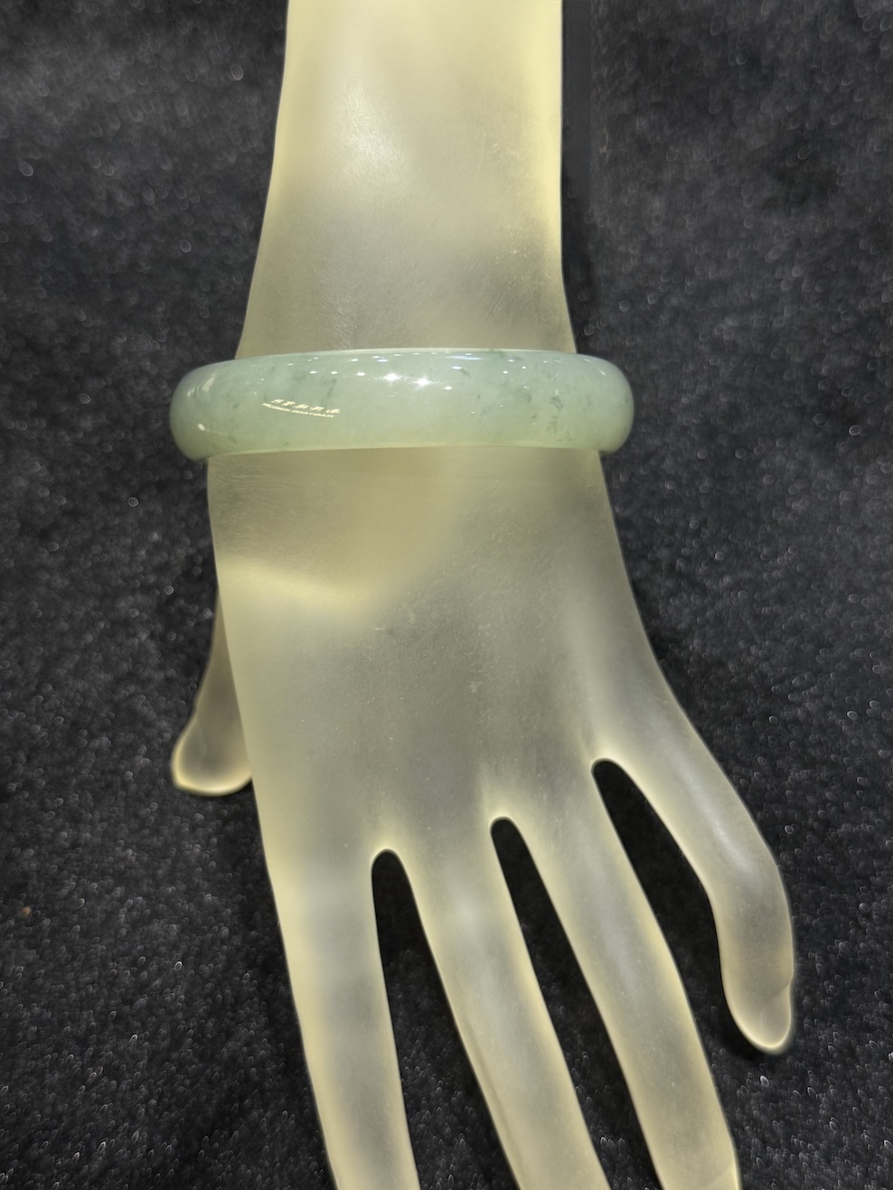 Translucent jadeite bangle in an icy, near-colourless body with a very faint green tint and small darker green flecks drifting through the ring